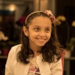 Descrição da imagem #PraCegoVer: Imagem no formato retangular, na horizontal. Nela, Sofia Crispim está sorrindo. Ela é uma menina morena, com longos cabelos castanhos, lisos. Na foto, Sofia veste um vestido com estampa cor de rosa e um casaquinho branco. Fim da descrição.