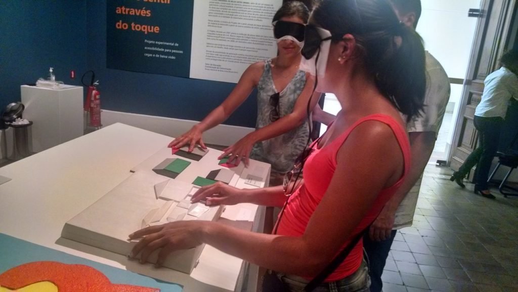 Descrição da imagem #PraCegoVer: A imagem está no formato retangular, na horizontal. Nela, duas jovens conhecem obras expostas pelo museu por meio das maquetes táteis. Elas estão acompanhadas de um guia e ambas têm os olhos vendados. Fim da descrição.