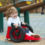 Descrição da imagem #PraCegoVer: A imagem está no formato retangular, na horizontal. Nela está a pequena Ana Paula em seu carrinho Fly Children. O carrinho é vermelho, tem duas rodas pretas e Ana está usando um vestido azul, estampado, uma blusa e meias branca. Ela está em um parque de diversões e sorri. Fim da descrição.