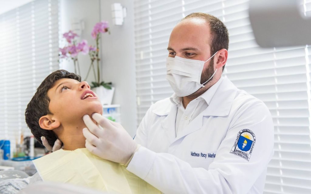 Descrição da imagem #pracegover: O dentista Matheus está em seu consultório odontológico, durante o atendimento a um paciente, que é um menino. O doutor está com jaleco branco e com uma máscara no rosto. O menino está deitado na cadeira de atendimento. Fim da descrição.