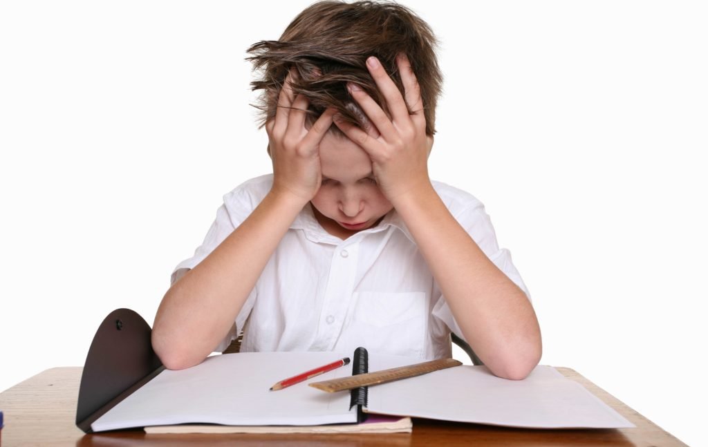 Descrição da imagem #PraCegoVer: Imagem no formato retangular, na horizontal. Nela, um menino está sentado em uma mesa, com um caderno aberto e alguns lápis espalhados pela mesa. O menino está com as duas mãos na cabeça, como se estivesse confuso com sua tarefa. Ele veste uma camisa branca. O fundo da imagem também é branco. Fim da descrição.