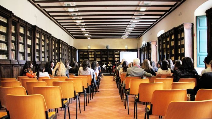 Descrição da imagem #PraCegoVer: A imagem está no formato retangular, na horizontal. Nela, é possível ver uma biblioteca, com dezenas de cadeiras de madeira. Algumas pessoas participam de uma reunião. Fim da descrição.