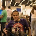 Descrição da imagem #PraCegoVer: A imagem está no formato retangular, na horizontal. Na imagem está o jovem Rafael, que também foi fotografado durante visita à Reatech, em um dos corredores do evento. Rafael está sentado em sua cadeira de rodas. Ele tem cabelos bem curtos, e é moreno. Ele usa uma camiseta do Capitão América. Fim da descrição.