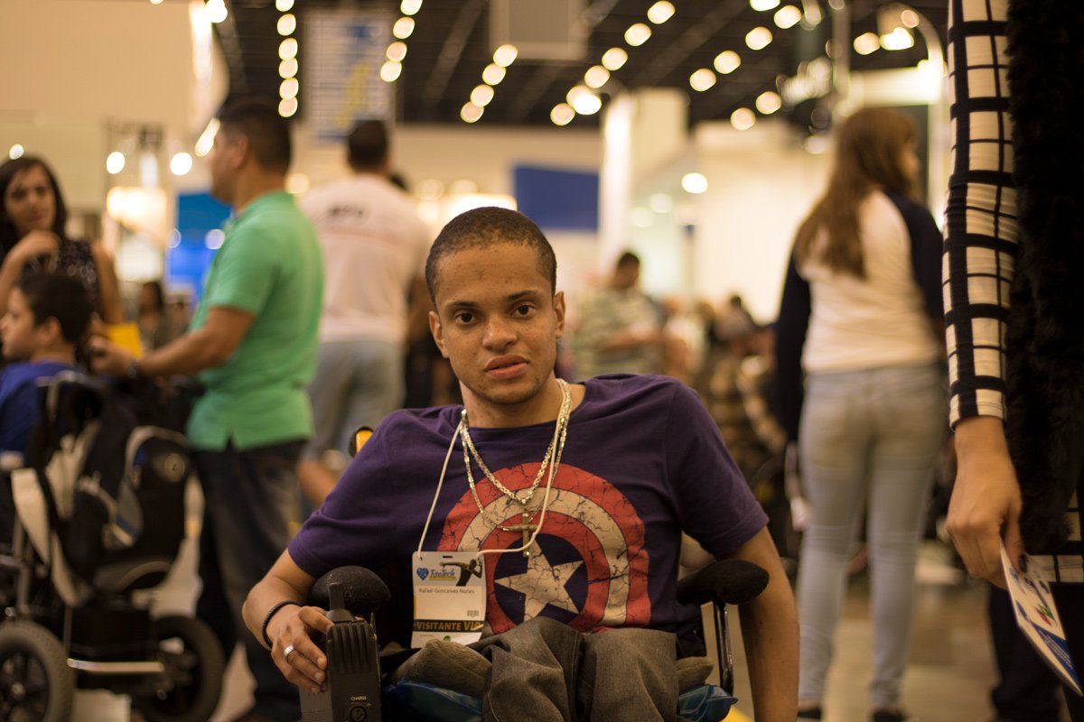 Descrição da imagem #PraCegoVer: A imagem está no formato retangular, na horizontal. Na imagem está o jovem Rafael, que também foi fotografado durante visita à Reatech, em um dos corredores do evento. Rafael está sentado em sua cadeira de rodas. Ele tem cabelos bem curtos, e é moreno. Ele usa uma camiseta do Capitão América. Fim da descrição.