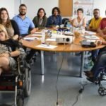 Descrição da imagem #PraCegoVer: Imagem no formato retangular, na horizontal. Nela estão as 11 pessoas que participaram da reunião preparatória das ações inclusivas no evento. Entre eles estão três cadeirantes. Todos estão sentados em volta de uma mesa oval de madeira. Fim da descrição.