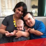 Descrição da imagem #PraCegoVer: Imagem no formato quadrado. Leo está acompanhado de sua esposa, Carolina, e de sua filha, a pequena Luísa, que é um lindo bebê. Carolina está vestindo uma blusa preta e um colar amarelo. Ela é uma mulher morena, e tem cabelos pretos, lisos e curtos. Leo está usando uma camiseta azul e segura a mão de sua filha. Luísa está vestindo um vestido preto com bolinhas brancas. Abaixo da imagem, foi inserida uma borda com formato irregular, na cor vermelha. Fim da descrição.