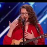 Descrição da imagem #PraCegoVer: A imagem está no formato retangular, na horizontal. Nela está a cantora surda Mandy Harvey. Ela está no palco e a foto foi feita durante sua conversa com os jurados. Ela tem um microfone à frente de seu corpo. Mandy é uma jovem morena, com cabelos castanhos, cacheados e longos. Ela usa óculos. Ela usa um vestido vermelho e está com seu cavaquinho. Fim da descrição.