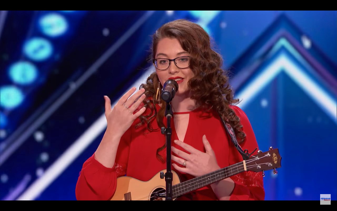 Descrição da imagem #PraCegoVer: A imagem está no formato retangular, na horizontal. Nela está a cantora surda Mandy Harvey. Ela está no palco e a foto foi feita durante sua conversa com os jurados. Ela tem um microfone à frente de seu corpo. Mandy é uma jovem morena, com cabelos castanhos, cacheados e longos. Ela usa óculos. Ela usa um vestido vermelho e está com seu cavaquinho. Fim da descrição.