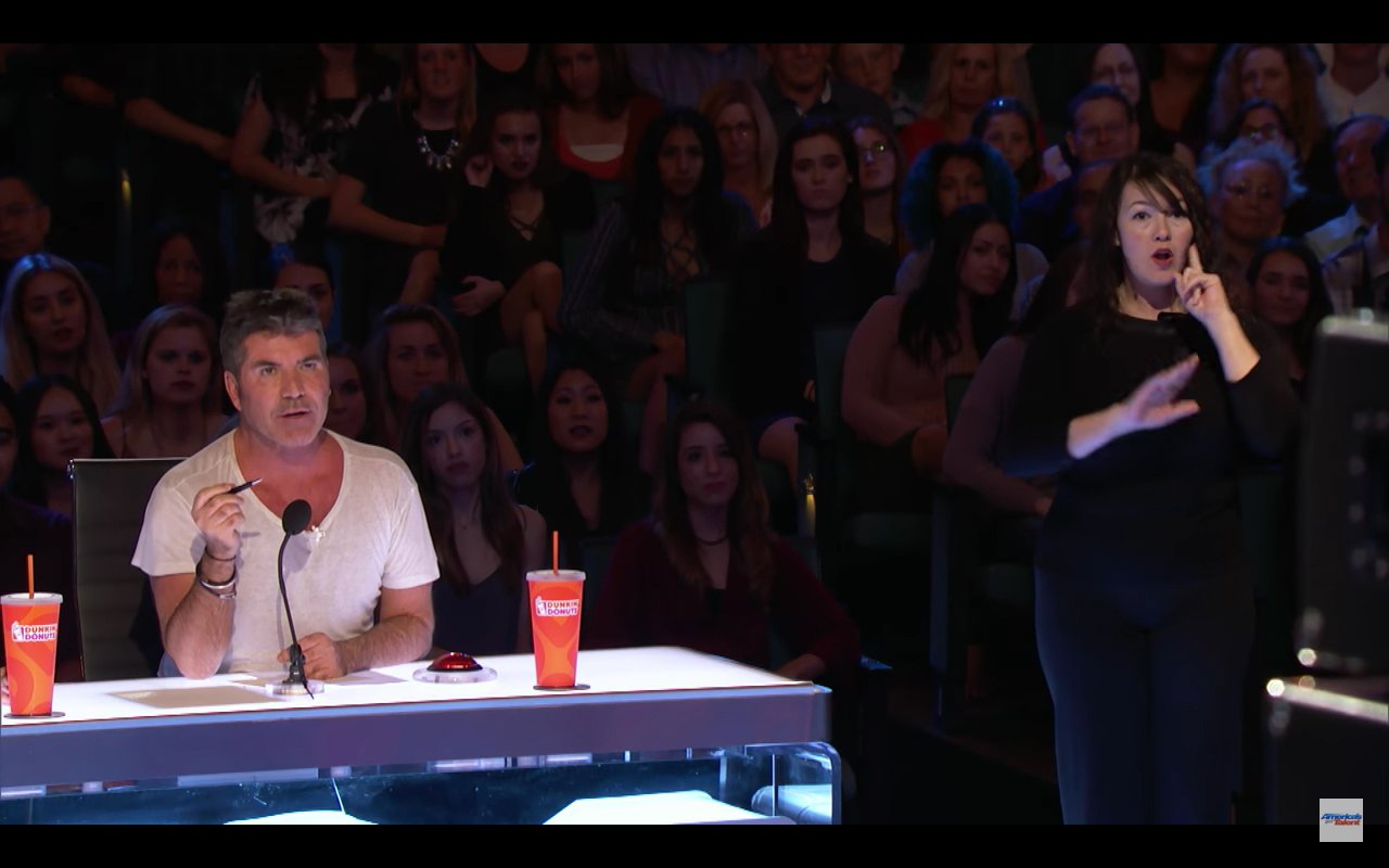 Descrição da imagem #PraCegoVer: A imagem está no formato retangular, na horizontal. Nela, ao fundo tem a platéia do programa American's Got Talent, e na frente, está sentado atrás de uma mesa, o jurado Simon Cowell usando uma camiseta branca. Ao lado, a intérprete da língua de sinais americana (ASL), Sarah, usando roupas pretas e fazendo um sinal. Fim da descrição.