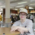 Descrição da imagem #PraCegoVer: A imagem está no formato retangular, na horizontal. Nela está a jovem Beatriz, funcionária do McDonald’s. Ela está sentada e ao fundo é possível ver a área de atendimento da lanchonete. Beatriz está uniformizada e com um boné da empresa. Ela usa óculos e está com um sorriso no canto da boca. Fim da descrição.