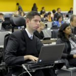 Descrição da imagem #PraCegoVer: Imagem no formato retangular, na horizontal. Nela temos o registro de uma das reuniões da convenção da ONU, em Nova Iorque. Na imagem temos, em primeiro plano, um homem com deficiência física, sentado em sua cadeira de rodas. Ele tem um computador portátil à sua frente. O homem usa camisa branca e um agasalho preto. Ao fundo, em segundo plano, vemos outras pessoas que participam da reunião. Fim da descrição.