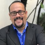 Descrição da imagem #PraCegoVer: A imagem está no formato retangular, na vertical. Nela está o educador Rafael Silva. Ele está próximo a uma parede branca. Ao fundo, do lado direito, é possível ver algumas árvores e folhagens, como uma praça. Rafael usa um terno cinza escuro, uma camisa azul royal e está com os braços cruzados. Ele está sorrindo. Fim da descrição.
