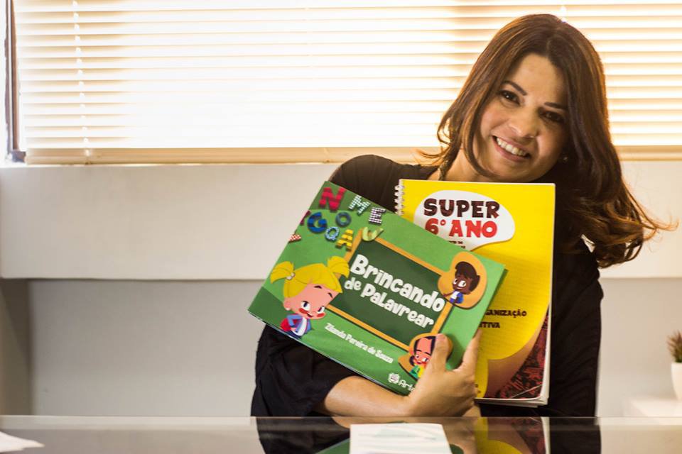 Descrição da imagem #PraCegoVer: Imagem no formato retangular, na horizontal. Nela, Zilanda Souza está sentada à frente de uma mesa, e segura alguns de seus livros: Brincando de Palavrear e o livro do programa de treino em funções executivas Super 6º Ano. Fim da descrição.