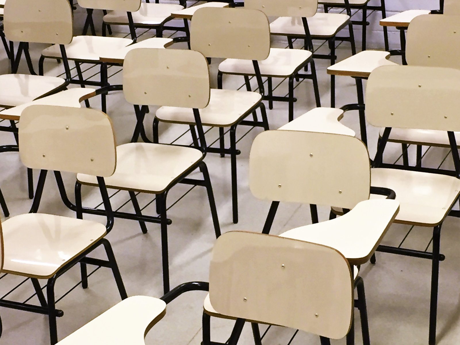 Descrição da imagem #PraCegoVer: Imagem no formato retangular, na horizontal. A Imagem é de uma sala de aula, com diversas carteiras na cor bege, vazias enfileiradas. Entre elas estão cadeiras com braços para destros e canhotos. Fim da descrição.