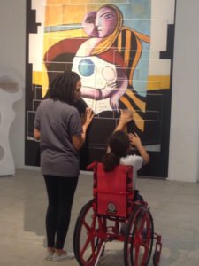 Descrição da imagem #PraCegoVer: Imagem no formato retangular, na vertical. Sofia está sentada em sua cadeira de rodas, tocando uma obra de arte do museu. Sofia está acompanhada de uma monitora do museu. Fim da descrição.