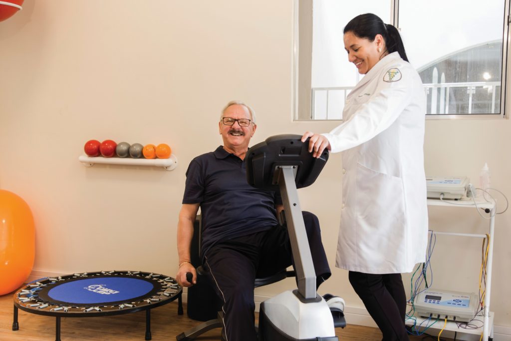 Descrição da imagem #PraCegoVer: Imagem no formato retangular, na horizontal. Um homem idoso está em uma sala de ginástica, realizando um exercício na bicicleta ergométrica. A seu lado está uma profissional, que está acompanhando seu exercício. Fim da descrição.