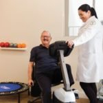 Descrição da imagem #PraCegoVer: Imagem no formato retangular, na horizontal. Um homem idoso está em uma sala de ginástica, realizando um exercício na bicicleta ergométrica. A seu lado está uma profissional, que está acompanhando seu exercício. Fim da descrição.