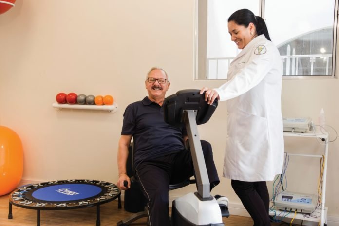 Descrição da imagem #PraCegoVer: Imagem no formato retangular, na horizontal. Um homem idoso está em uma sala de ginástica, realizando um exercício na bicicleta ergométrica. A seu lado está uma profissional, que está acompanhando seu exercício. Fim da descrição.