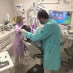 Descrição da imagem #PraCegoVer: A imagem está no formato retangular, na vertical. Nela, o doutor Matheus está em seu consultório odontológico, durante o atendimento a um paciente. O doutor recebe o auxílio de uma assistente. A assistente usa um jaleco lilás e o doutor Matheus usa um jaleco verde. Ambos estão com calça branca. Fim da descrição.
