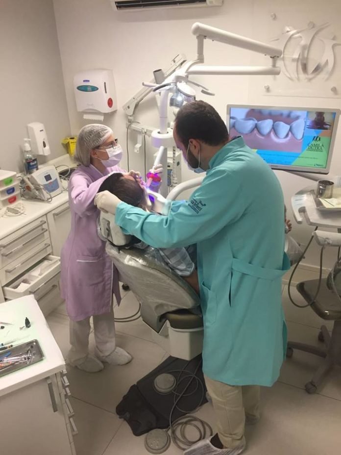 Descrição da imagem #PraCegoVer: A imagem está no formato retangular, na vertical. Nela, o doutor Matheus está em seu consultório odontológico, durante o atendimento a um paciente. O doutor recebe o auxílio de uma assistente. A assistente usa um jaleco lilás e o doutor Matheus usa um jaleco verde. Ambos estão com calça branca. Fim da descrição.