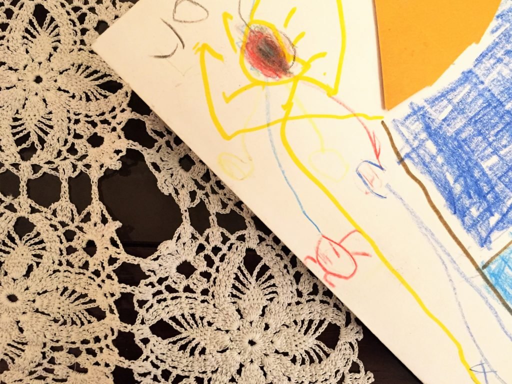 Descrição da imagem #PraCegoVer: Imagem no formato retangular, na horizontal. A imagem tem uma composição, com um pedaço de uma folha de sulfite, com um desenho infantil, sem uma definição clara. O desenho está em cima de uma mesa. Abaixo da folha com o desenho está uma toalha branca de crochê bordada. Fim da descrição.