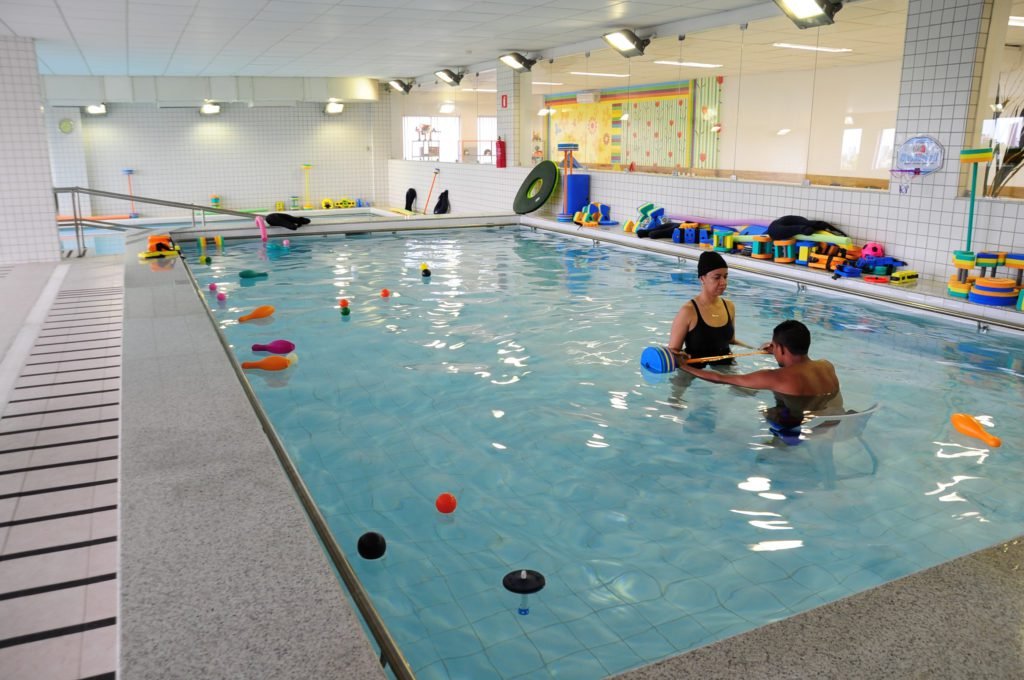 Descrição da imagem #PraCegoVer: Imagem no formato retangular, na horizontal. Uma das piscinas do centro de reabilitação, que já está em funcionamento. Nela vemos uma sessão de hidroterapia, realizada por uma profissional em um rapaz jovem, que está na piscina, sentado em uma cadeira. Fim da descrição.