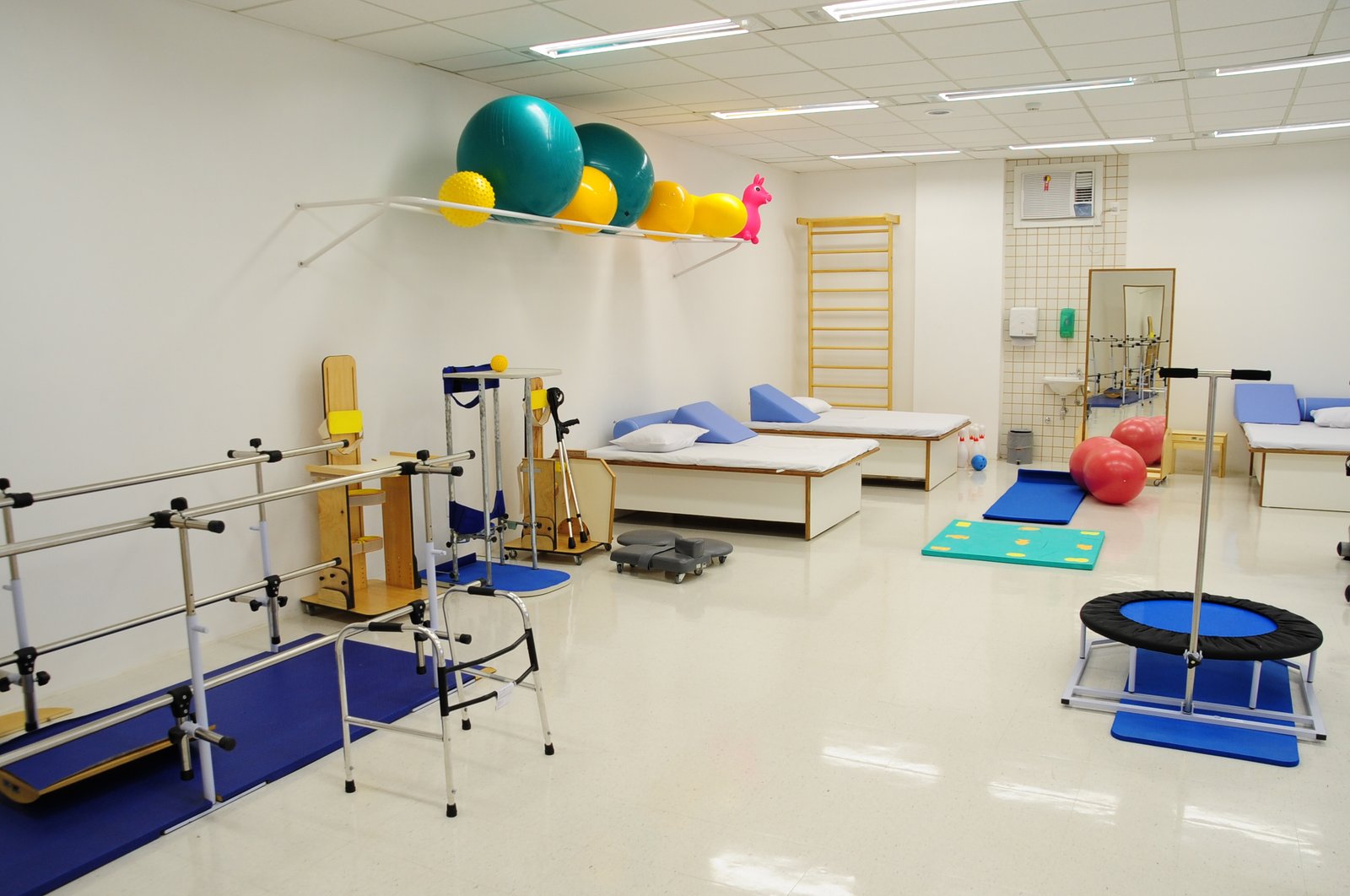 Descrição da imagem #PraCegoVer: Imagem no formato retangular, na horizontal. A sala de fisioterapia do centro de reabilitação. Nela vemos bolas, um jumper, duas áreas de atendimento, um andador e barras auxiliares para treino de marcha, entre outros equipamentos. Fim da descrição.