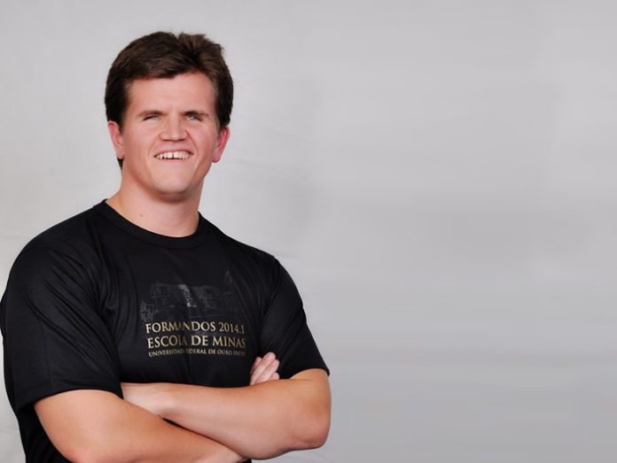 Descrição da imagem #PraCegoVer: Imagem no formato retangular, na horizontal. O jovem Felipe Rigoni, posa para a foto. Ele está no canto esquerdo da imagem. Felipe usa uma camiseta preta, em que está escrito, em letras douradas: Formandos 2014 Escola de Minas. Felipe é um rapaz moreno, de pele clara e cabelos castanhos escuros, curtos. Ele está com os braços cruzados e sorri. Fim da descrição.