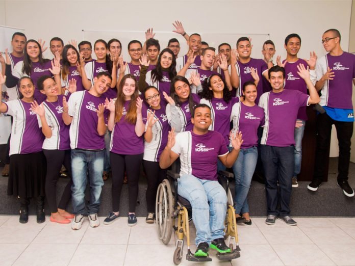 Descrição da imagem #pracegover: Um grupo com muitos jovens, todos uniformizados, com a camiseta do IOS. Entre os jovens, vemos um na frente, em destaque, que tem deficiência física. Ele está sentado em sua cadeira de rodas. Fim da descrição.