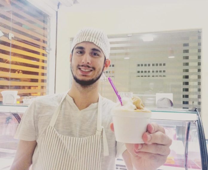 Descrição da imagem #PraCegoVer: Imagem no formato retangular, na horizontal. O empresário Breno Nunes, dono da gelateria il Sordo, posa em seu estabelecimento. Ele está segurando um copo de seu gelato. Breno é um rapaz moreno, de pele clara. Ele tem barba e usa uma bandana branca na cabeça. Breno veste camisa e avental brancos. Fim da descrição.