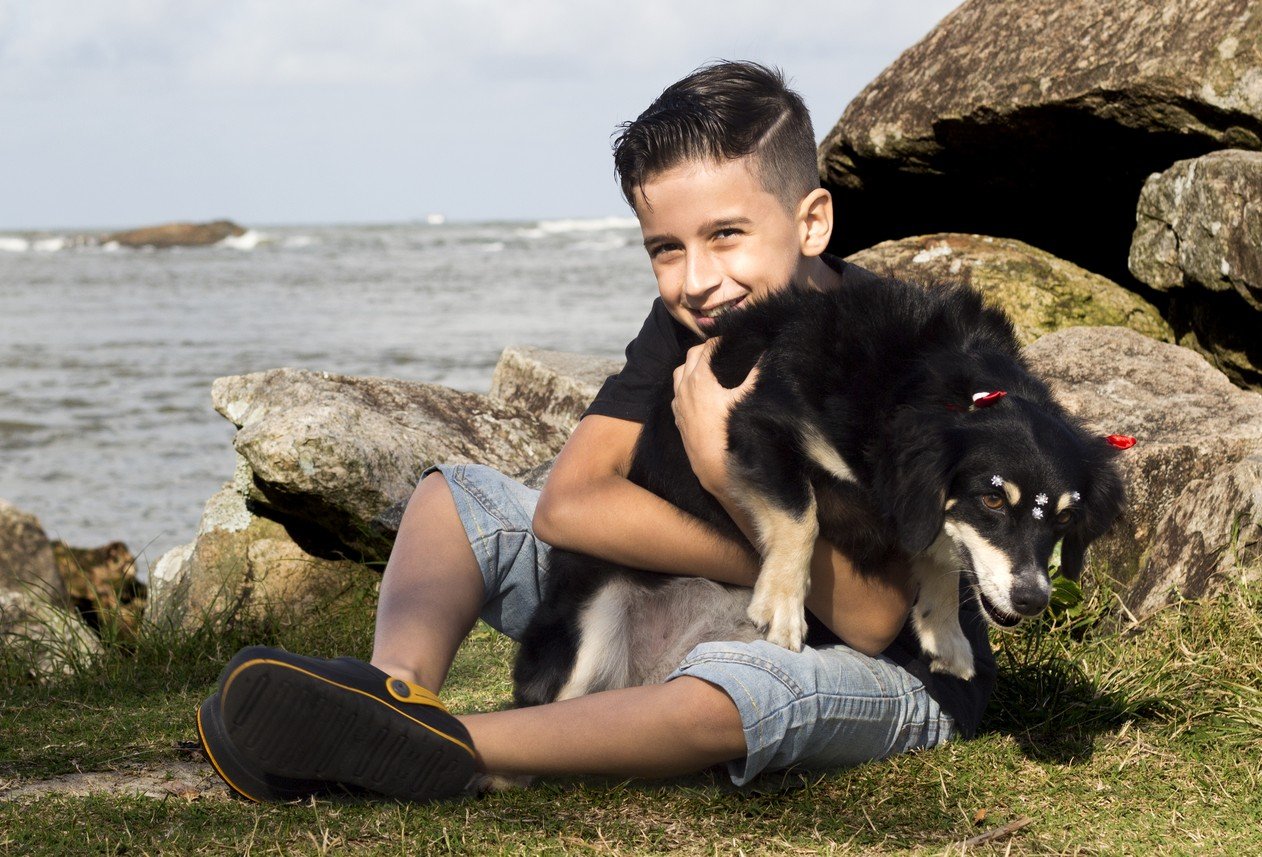 Descrição da imagem #PraCegoVer: Imagem no formato retangular, na horizontal. Yuri está em uma praia com a cachorrinha Mandy no colo. Ele está sentado no chão. Yuri é um menino moreno, com pele clara e cabelos castanhos e curtos. Mandy é uma cachorrinha vira-lata, de pelos pretos e detalhes na cor branca. Ao fundo vemos o mar. Fim da descrição.