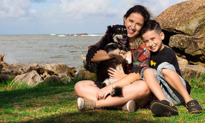 Descrição da imagem #PraCegoVer: Imagem no formato retangular, na horizontal. Yuri e sua mãe estão sentados no chão. Eles estão em uma praia com a cachorrinha Mandy, que está no colo de Denise. O menino está sentado ao lado delas. Yuri é um menino moreno, com pele clara e cabelos castanhos e curtos. Sua mãe também é morena, com pele clara, e tem cabelos castanhos, lisos e compridos. Mandy é uma cachorrinha vira-lata, de pelos pretos e pequenos detalhes na cor branca. Yuri e sua mãe estão sorrindo. Ao fundo vemos o mar e ao lado algumas pedras grandes. Fim da descrição.