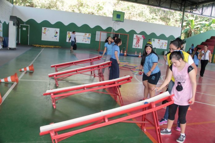 Descrição da imagem #PraCegoVer: Imagem no formato retangular, na horizontal. Jovens com deficiência intelectual participam de uma partida de bocha adaptada. Vemos quatro meninas que jogam suas bolas nas canaletas, que garantem a adaptação da modalidade. Elas estão em uma quadra poliesportiva. Fim da descrição. 