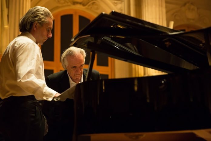 Descrição da imagem #PraCegoVer: Imagem no formato retangular, na horizontal. O ator Alexandre Nero e o maestro João Carlos Martins durante as gravações do filme João, o maestro. Alexandre e o maestro estão ao lado de um grande piano, de calda. Alexandre está caracterizado como o maestro. Ele usa uma calça preta e uma camisa branca. Ele está com os cabelos compridos e brancos. João Carlos Martins usa terno preto e camisa branca. Fim da descrição.
