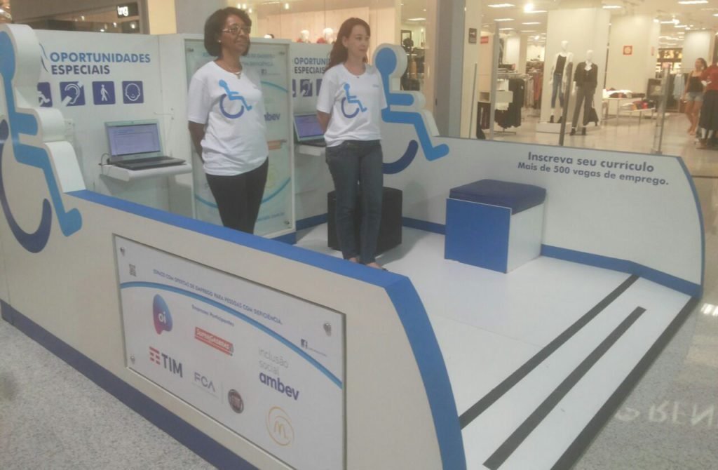 Descrição da imagem #PraCegoVer: A imagem está no formato retangular, na horizontal. Nela é possível ver o estande do Oportunidades Especiais instalado em um corredor de shopping center. Duas monitoras do projeto estão no local. Fim da descrição.