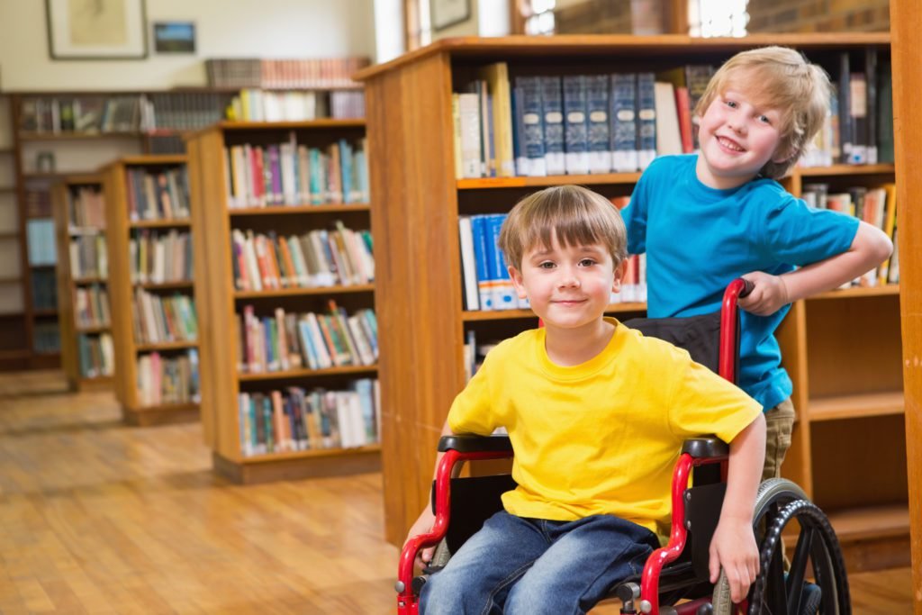 Descrição da imagem #PraCegoVer: Imagem no formato retangular, na horizontal. Dois alunos estão em uma biblioteca de escola. Um dos meninos é cadeirante e está em sua cadeira de rodas, o outro menino está em pé, atrás da cadeira. Os meninos são loiros. O menino cadeirante usa camiseta amarela e calça jeans. O menino que está em pé usa uma camiseta azul. Fim da descrição.