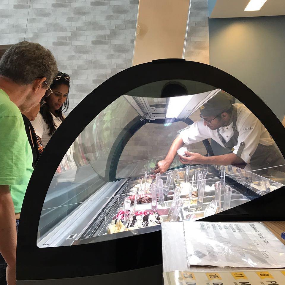 Descrição da imagem #PraCegoVer: Imagem no formato retangular, na horizontal. O balcão de atendimento da gelateria. Um atendente surdo está servindo alguns clientes. Ele está pegando sorvete na vitrine. Fim da descrição.
