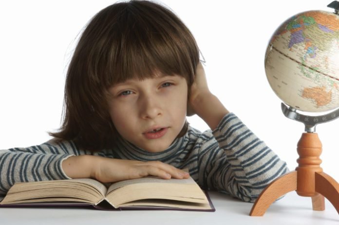 Descrição da imagem #PraCegoVer: Um menino está sentado e à sua frente está um livro aberto e um globo terrestre. O menino é loiro e tem cabelos lisos. Ele está com a mão direita sobre o livro e a esquerda apoiada em sua cabeça. Ele parece está pensando em algo distante. Fim da descrição.