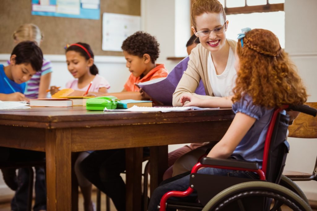 Descrição da imagem #pracegover: foto de uma sala de aula. Uma educadora está sentada ao lado de uma menina cadeirante. A menina está de costas e olha para a professora. Na mesma mesa estão outras duas crianças, uma menina e um menino. Todos estão felizes. Fim da descrição.