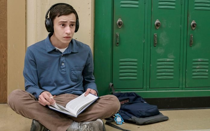 Descrição da imagem #PraCegoVer: Imagem no formato retangular, na horizontal. O personagem Sam, protagonista da série, está sentado no corredor da escola. Ele está sentado no chão, ao lado do armário escolar, verde. Sam veste calça na cor marrom e camisa jeans. Ele está com fones de ouvido e com um caderno aberto em seu colo. Fim da descrição.
