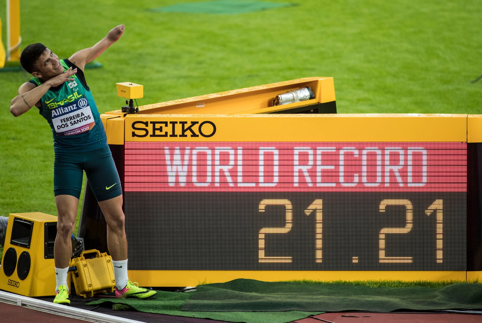 Descrição da imagem #PraCegoVer: Imagem no formato retangular, na horizontal. O atleta Petrúcio Ferreira após conquistar medalha de ouro e quebrar recorde dos 200 metros no Mundial de Londres. Petrúcio está ao lado da placa que anuncia a quebra de recorde. Ele imita o atleta Usain Bolt, que atualmente é o corredor mais rápido do mundo, fazendo a mesma posição, como se estivesse lançando uma flecha com os braços. Fim da descrição.