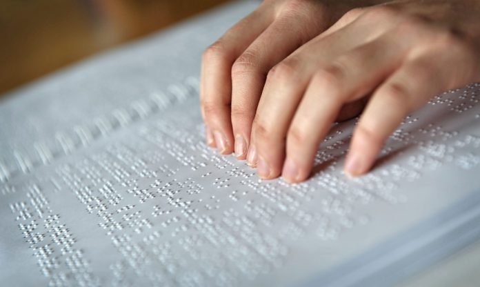 Descrição da imagem #pracegover: Foto de dedos fazendo a leitura em braile em um documento impresso. Fim da descrição.