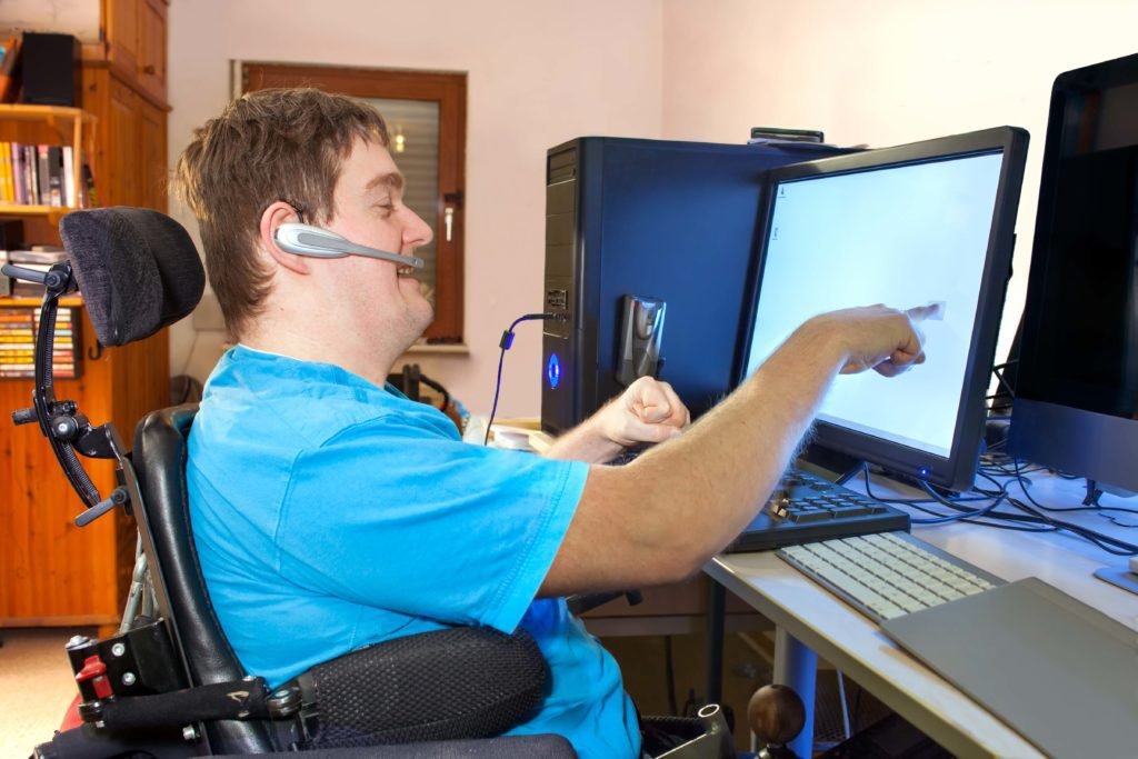 Descrição de Imagem #pracegover: Um rapaz com deficiência está em sua mesa de trabalho. Ele é cadeirante e está usando headfone e computador. O rapaz é moreno e tem cabelos castanhos, curtos. Ele está vestindo uma camiseta azul. Fim da descrição.