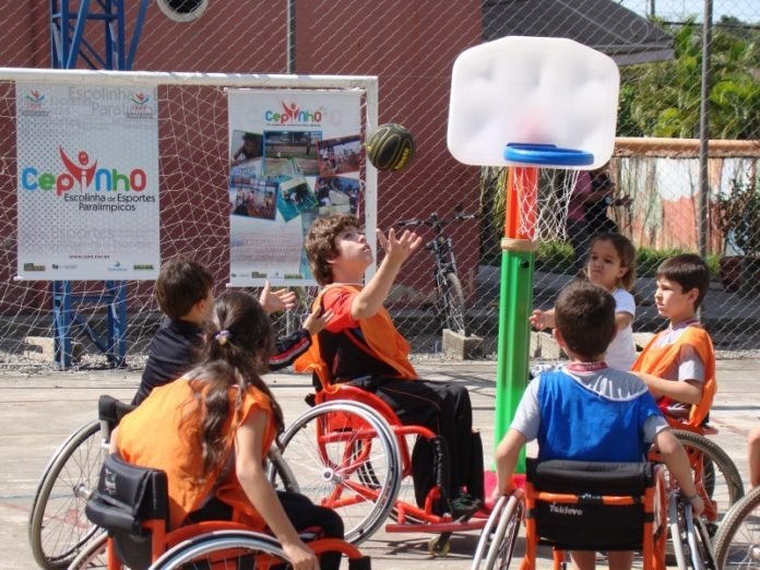 Descrição da imagem #PraCegoVer: Imagem no formato retangular, na horizontal. Crianças estão jogando basquete adaptado em uma quadra. Elas estão em suas cadeiras de rodas. Fim da descrição.