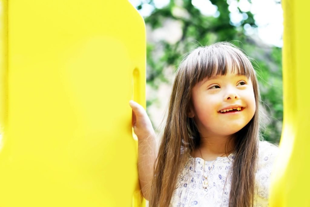 Descrição da imagem #PraCegoVer: Imagem no formato retangular, na horizontal. Uma menina com síndrome de Down está sorrindo. Ela está em um parque. A menina tem aproximadamente cinco anos de idade, ela é morena, tem a pele clara e cabelos castanhos, lisos e compridos. Ela está sorrindo. Fim da descrição.