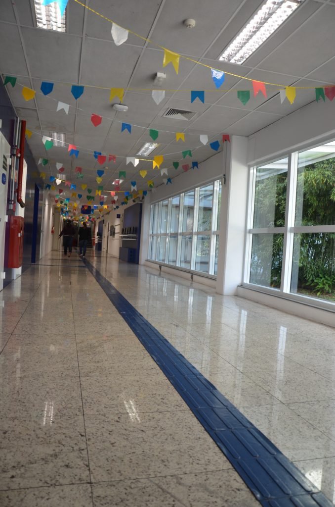 Descrição da imagem #PraCegoVer: Imagem no formato retangular, na vertical. O corredor da sede da TIM. O corredor é largo e bem iluminado. Ele tem piso podotátil e está decorado com bandeirinhas coloridas de festa junina. Fim da descrição.