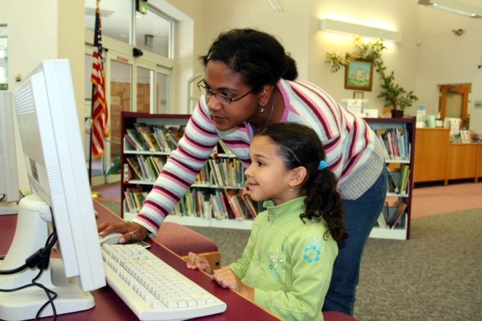 Descrição da imagem #pratodosverem: Em um ambiente escolar, uma aluna recebe auxílio de uma educadora. Elas estão à frente de um computador. Fim da descrição.