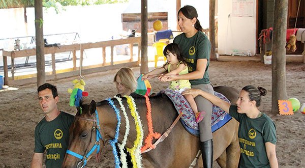 Descrição da imagem #PraCegoVer: Imagem no formato retangular, na horizontal. Uma sessão de fisioterapia na hípica Santa Terezinha. A equoterapeuta Eliane está montada no cavalo, realizando o atendimento a uma menina que tem deficiência intelectual. A menina está sentada na frente da profissional. Um homem conduz o cavalo e duas profissionais dão assistência de solo à equoterapeuta. O cavalo é marrom e está enfeitado com laços e fitas. A menina usa uma camiseta amarela e bermuda branca. Os profissionais usam uma camiseta verde, uniforme da hípica. Fim da descrição..