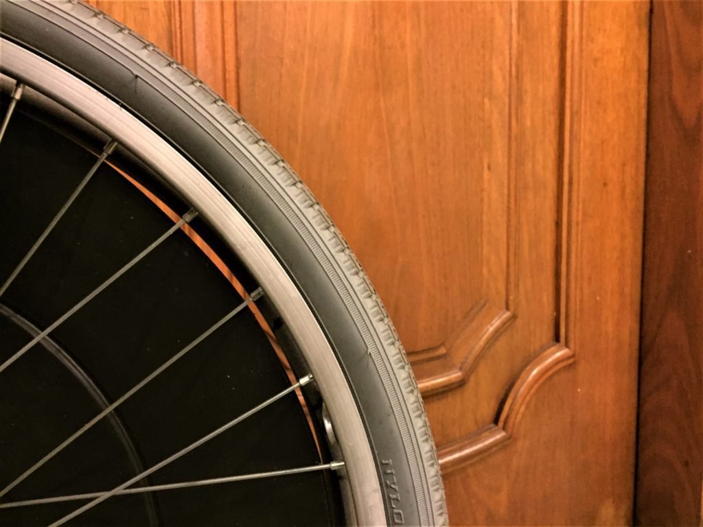 Descrição da imagem #pracegover: Em primeiro plano, uma roda de uma cadeira de rodas, e em segundo plano, uma porta de madeira, com detalhes. Fim da descrição.