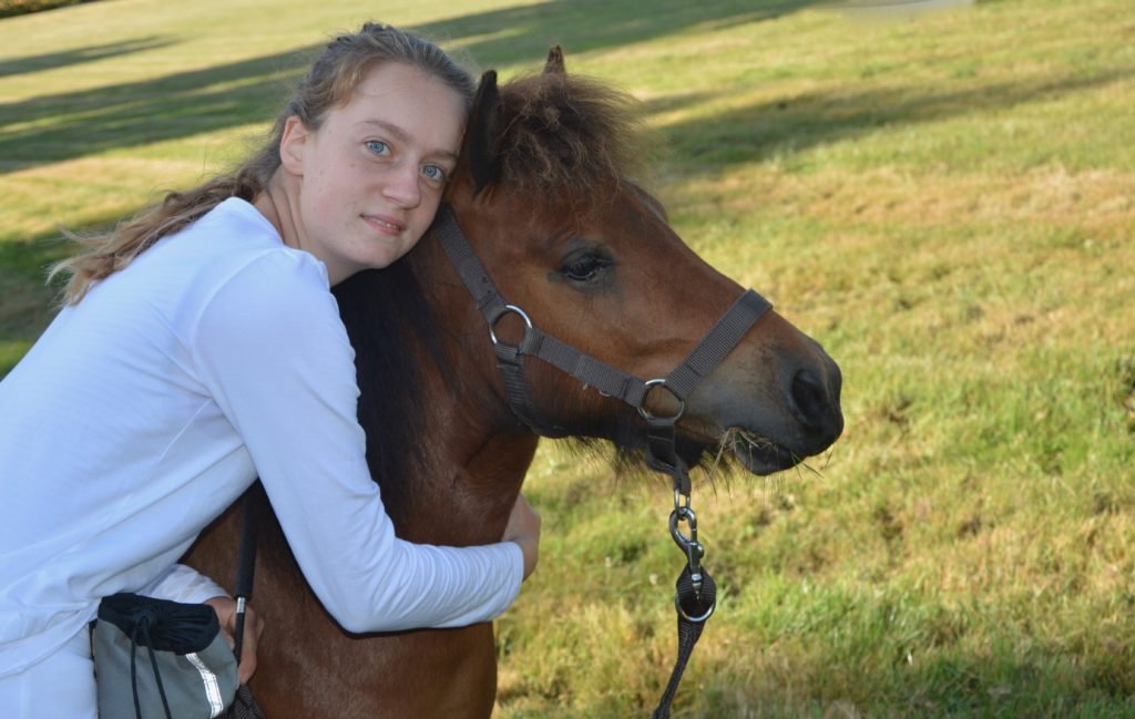Descrição da imagem #pracegover: Uma moça está abraçada a um cavalo. Ela é branca e tem cabelos castanhos. Ela usa uma roupa branca. O cavalo é marrom e está de lado. Ao fundo é possível ver um gramado verde. Fim da descrição.
